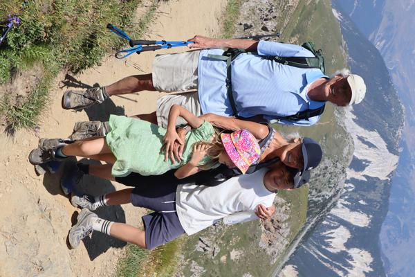 A family of four, including two adults and two children, poses happily on a sunny mountain trail